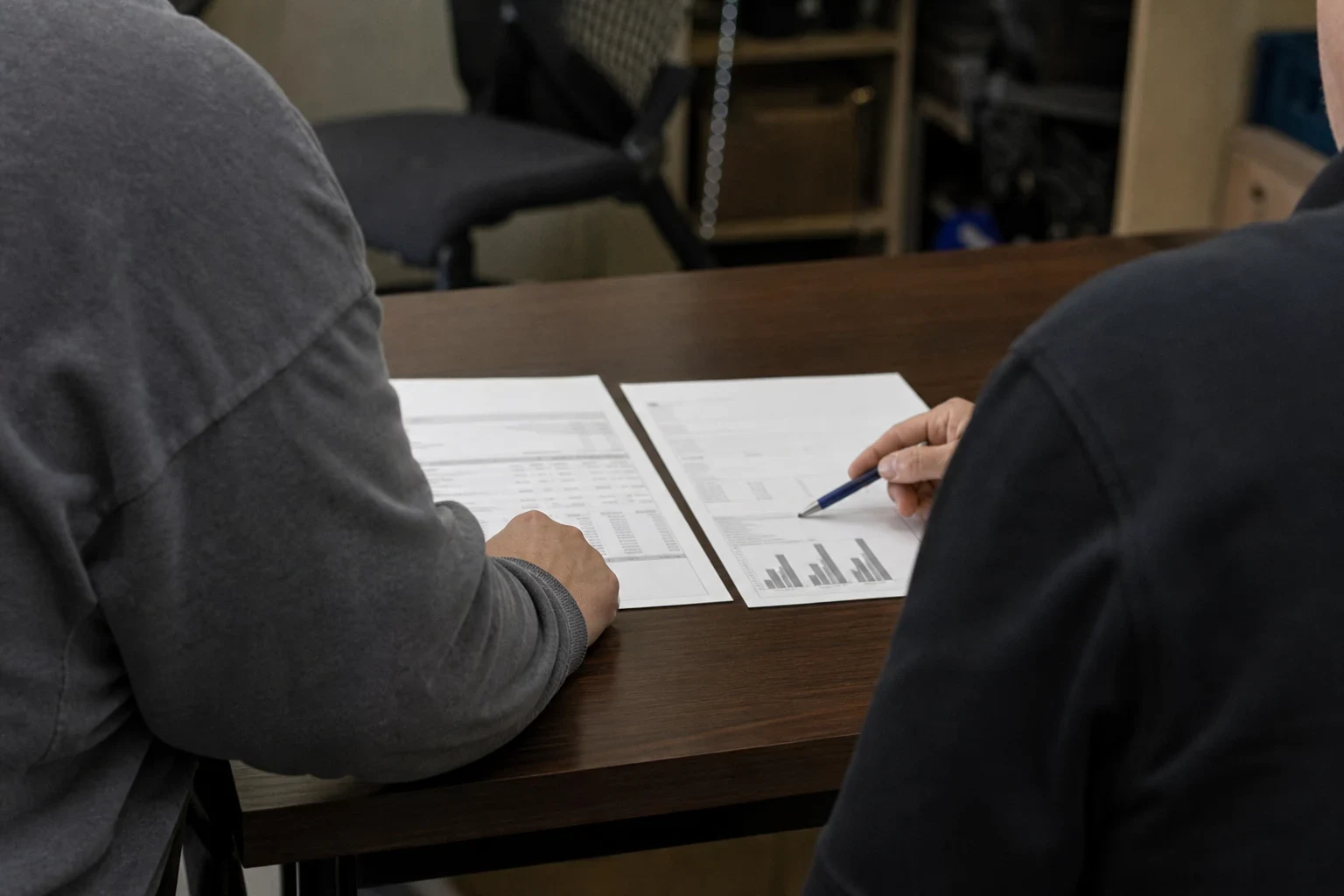 Two people at a dark wood office desk reviewing two printed documents side by side, one pointing with a pen at a page with a bar chart