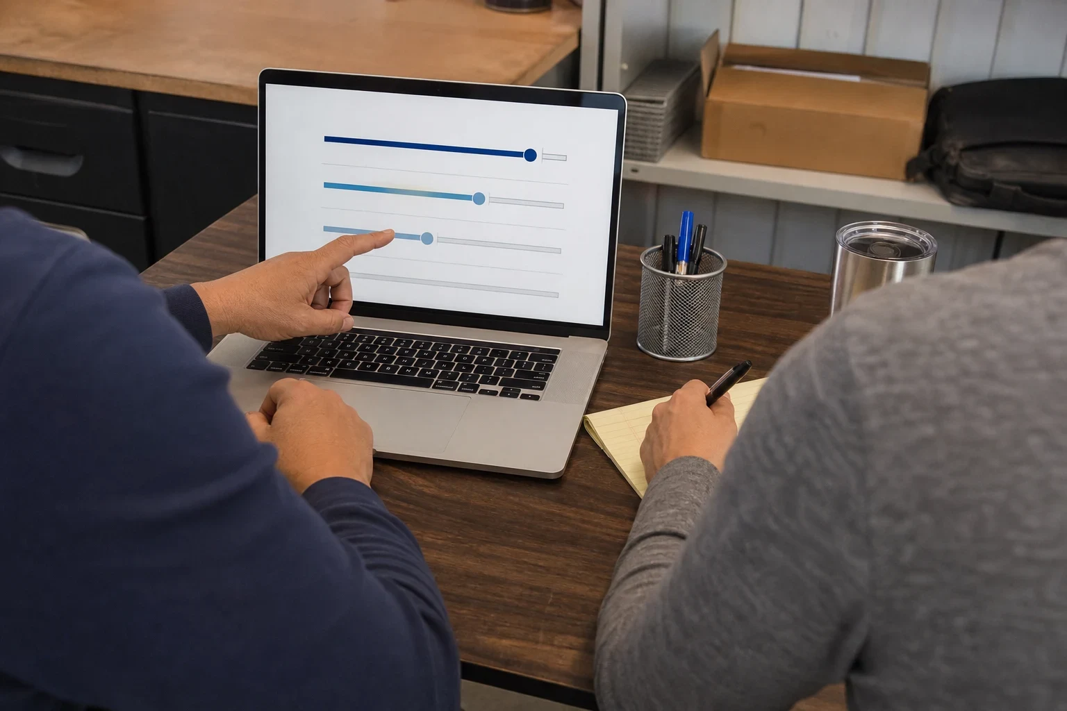 Over-the-shoulder view of an agent pointing at slider options on a laptop while a client takes notes on a yellow legal pad at an office desk